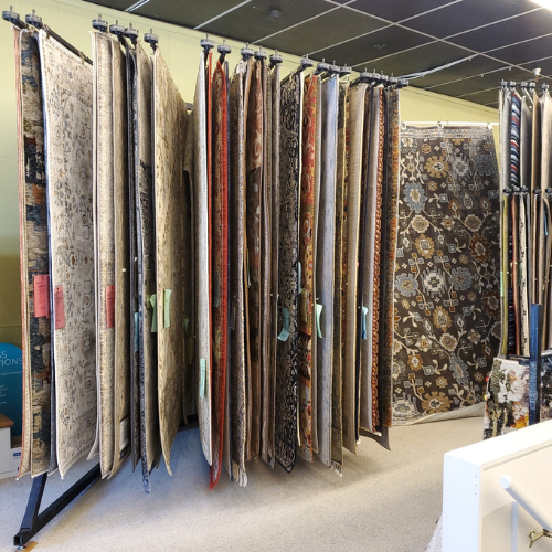Rows of hanging area rugs and patterned textiles on display in a showroom, with a large patterned rug on the wall right behind stairs